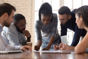 Focused African American team leader leaned over table, writing down project ideas, editing documentation at brainstorming business meeting with diverse partners or clients at office.