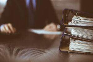 A man in a black suit with a black tie writes on a piece of paper. In the corner are three stacked binders with paper in them. 